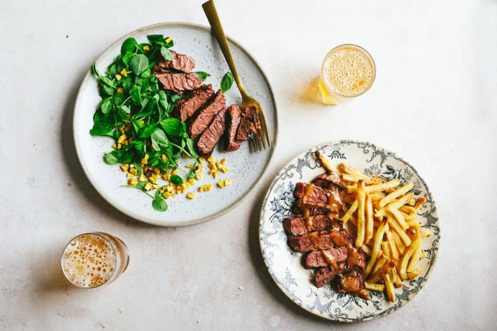 Two plated-up mycelium steak meals, one with green leaves and sweet corn, the other frites — shot from above with drinks on white surface.