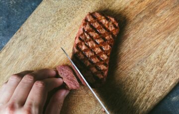 Close-up of griddled-brown mycelium steak being cut with a knife on wood chopping board to show reddish-pink interior texture similar to beef.