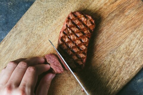 Close-up of griddled-brown mycelium steak being cut with a knife on wood chopping board to show reddish-pink interior texture similar to beef.