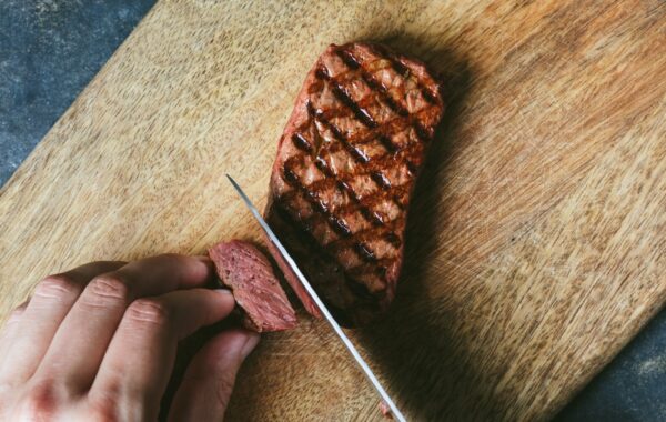 Close-up of griddled-brown mycelium steak being cut with a knife on wood chopping board to show reddish-pink interior texture similar to beef.