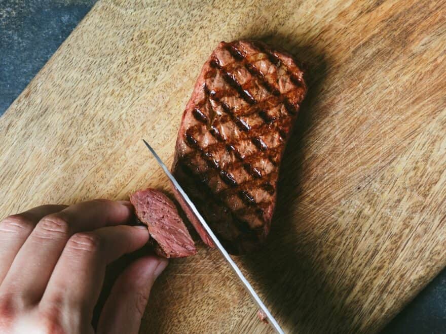 Close-up of griddled-brown mycelium steak being cut with a knife on wood chopping board to show reddish-pink interior texture similar to beef.