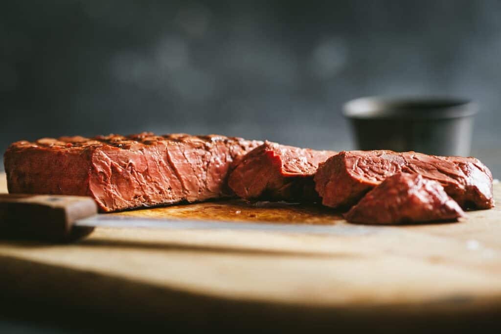 Close-up of cooked and cut mycelium steak on wood chopping board, showing textured reddish-pink interior.