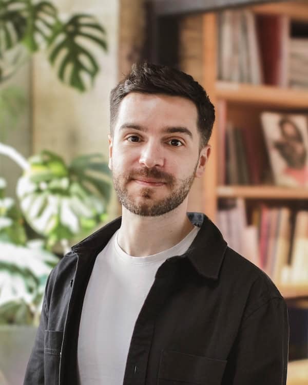 James Addison smiling, mouth closed, to camera, wearing a black 'shacket' over white T-shirt; out-of-focus large green plants and bookcases behind.