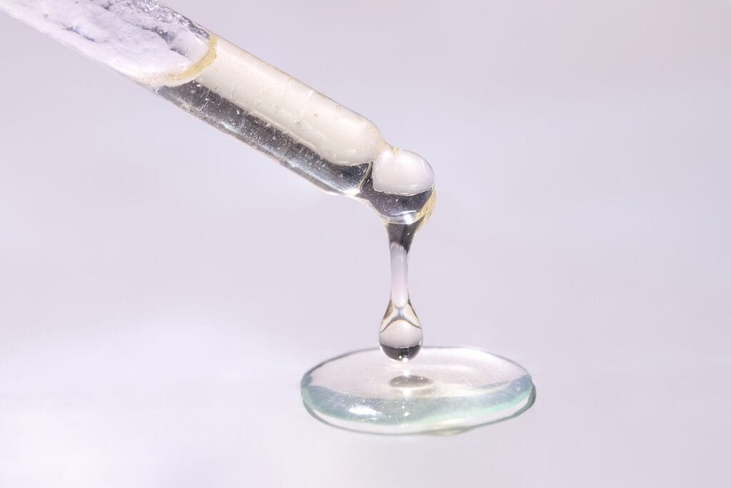 Close-up of droplets of mostly clear liquid fat hanging from end of pipette and pooling below, against pinkish white backdrop.