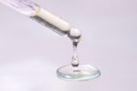Close-up of droplets of mostly clear liquid fat hanging from end of pipette and pooling below, against pinkish white backdrop.