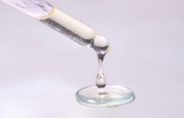 Close-up of droplets of mostly clear liquid fat hanging from end of pipette and pooling below, against pinkish white backdrop.