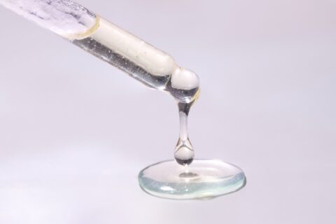 Close-up of droplets of mostly clear liquid fat hanging from end of pipette and pooling below, against pinkish white backdrop.
