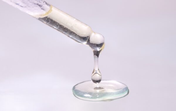 Close-up of droplets of mostly clear liquid fat hanging from end of pipette and pooling below, against pinkish white backdrop.
