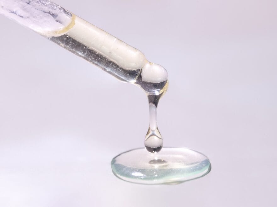Close-up of droplets of mostly clear liquid fat hanging from end of pipette and pooling below, against pinkish white backdrop.