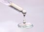 Close-up of droplets of mostly clear liquid fat hanging from end of pipette and pooling below, against pinkish white backdrop.