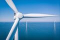 Offshore wind farm pictured against blue sea and sky, with one turbine in close-up in the foreground and two others shown in full at a distance.