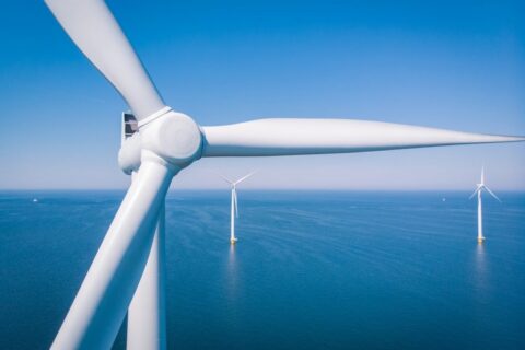 Offshore wind farm pictured against blue sea and sky, with one turbine in close-up in the foreground and two others shown in full at a distance.