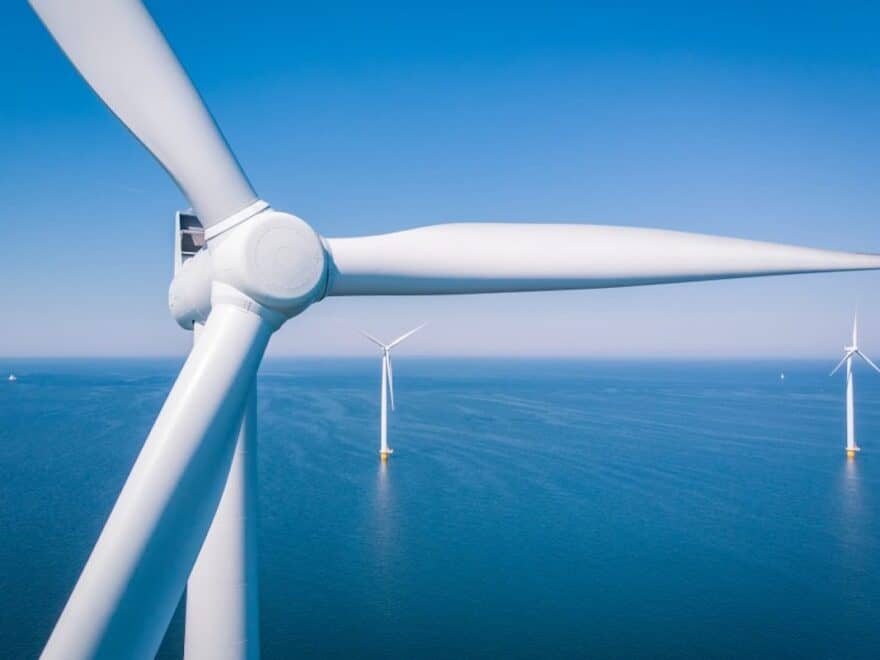 Offshore wind farm pictured against blue sea and sky, with one turbine in close-up in the foreground and two others shown in full at a distance.