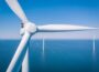 Offshore wind farm pictured against blue sea and sky, with one turbine in close-up in the foreground and two others shown in full at a distance.