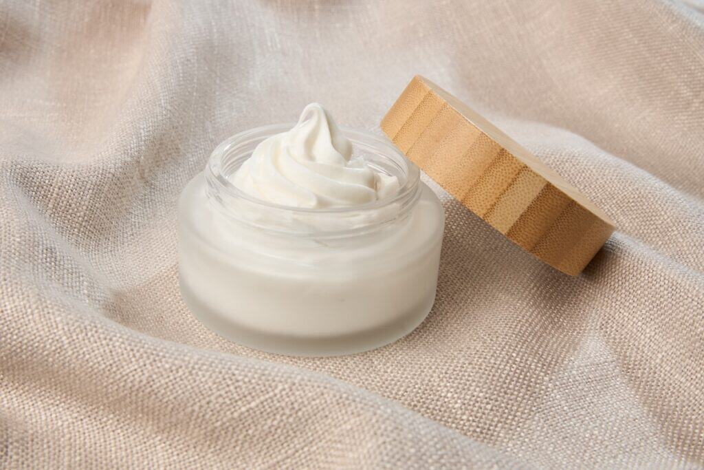 Open glass pot of bright white fat, looking like whipped cream, with peak sticking up and wooden lid off to the side, on wavy biscuit-coloured cloth.