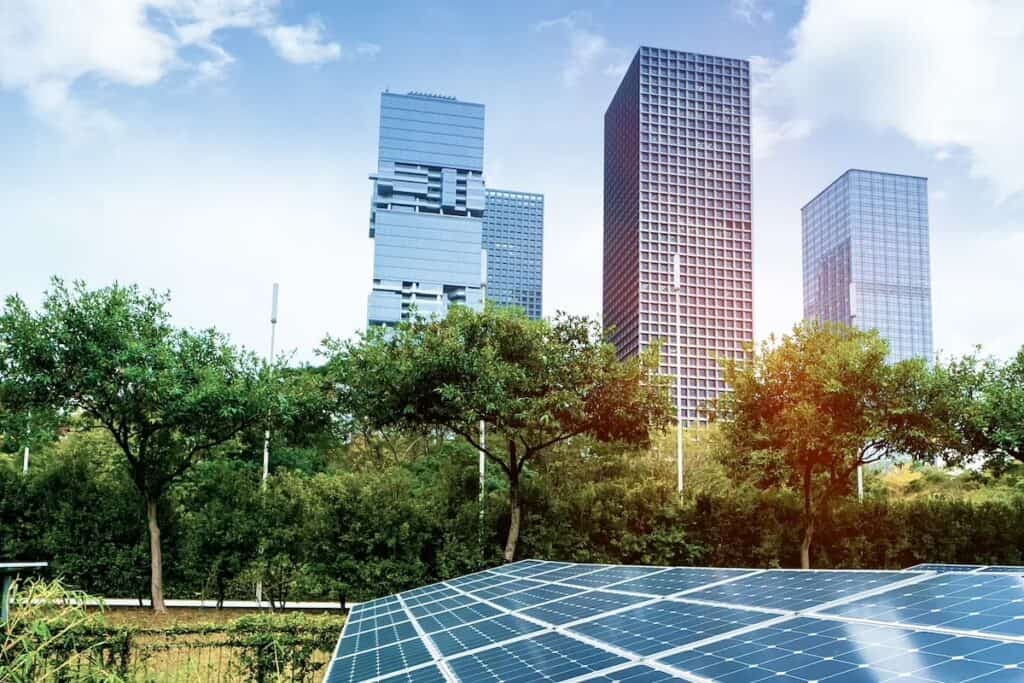 Colour filtered image of ground-level solar PV panels in foreground, with high-rise cityscape behind, plus trees and greenery between.