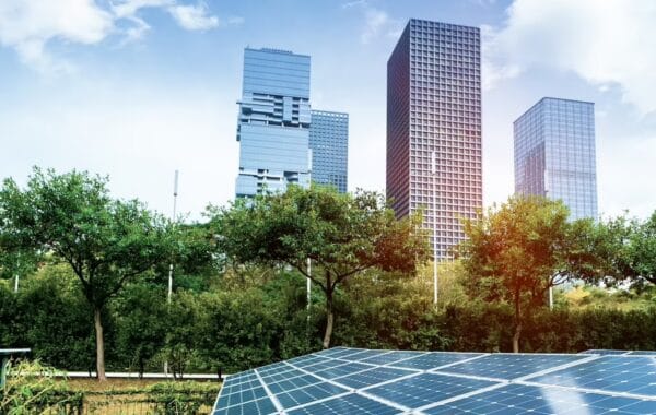 Colour filtered image of ground-level solar PV panels in foreground, with high-rise cityscape behind, plus trees and greenery between.