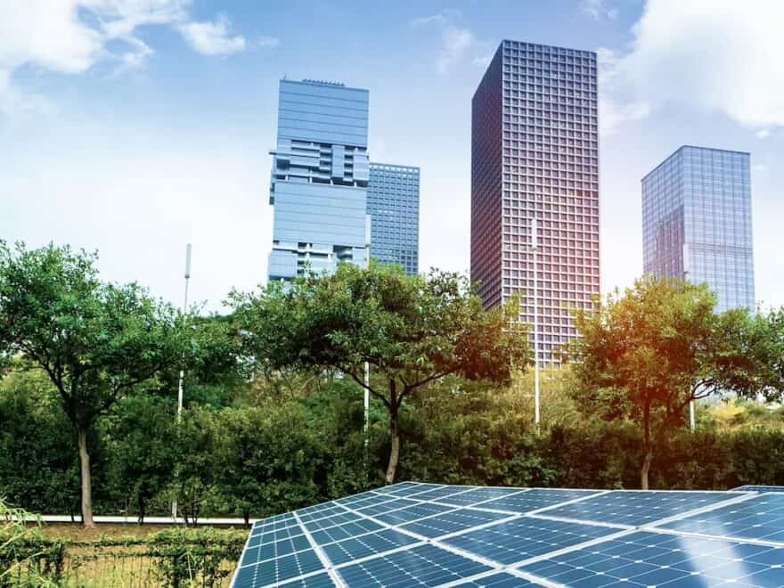 Colour filtered image of ground-level solar PV panels in foreground, with high-rise cityscape behind, plus trees and greenery between.