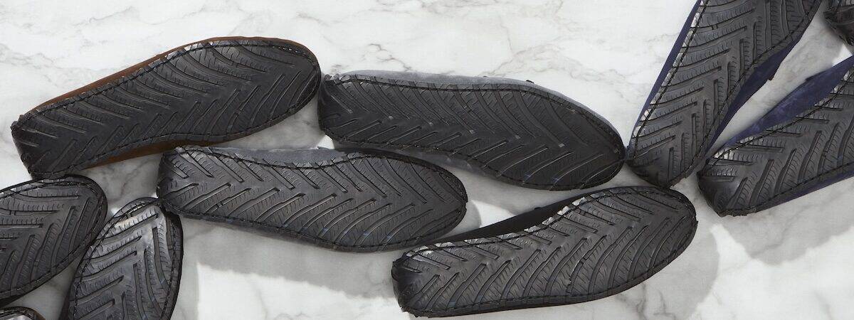 Multiple pairs of loafers, face-down snaking bottom L to top R, black upcycled-tyre soles with herringbone tread, on white and grey marble.