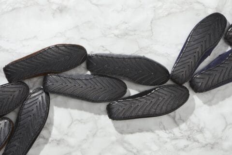 Multiple pairs of loafers, face-down snaking bottom L to top R, black upcycled-tyre soles with herringbone tread, on white and grey marble.