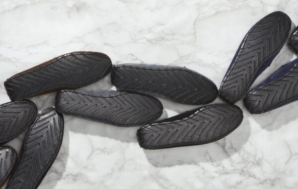 Multiple pairs of loafers, face-down snaking bottom L to top R, black upcycled-tyre soles with herringbone tread, on white and grey marble.