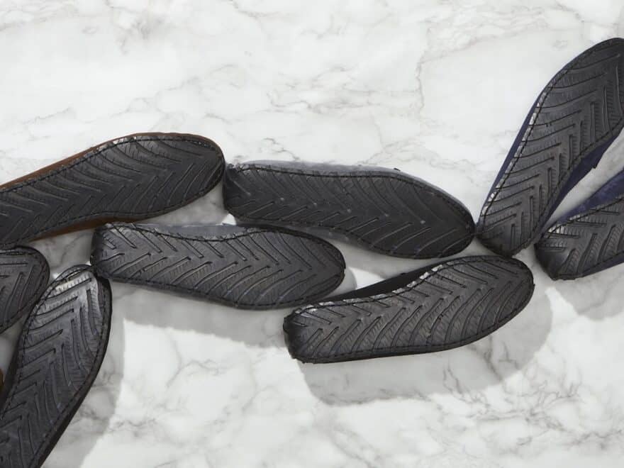 Multiple pairs of loafers, face-down snaking bottom L to top R, black upcycled-tyre soles with herringbone tread, on white and grey marble.