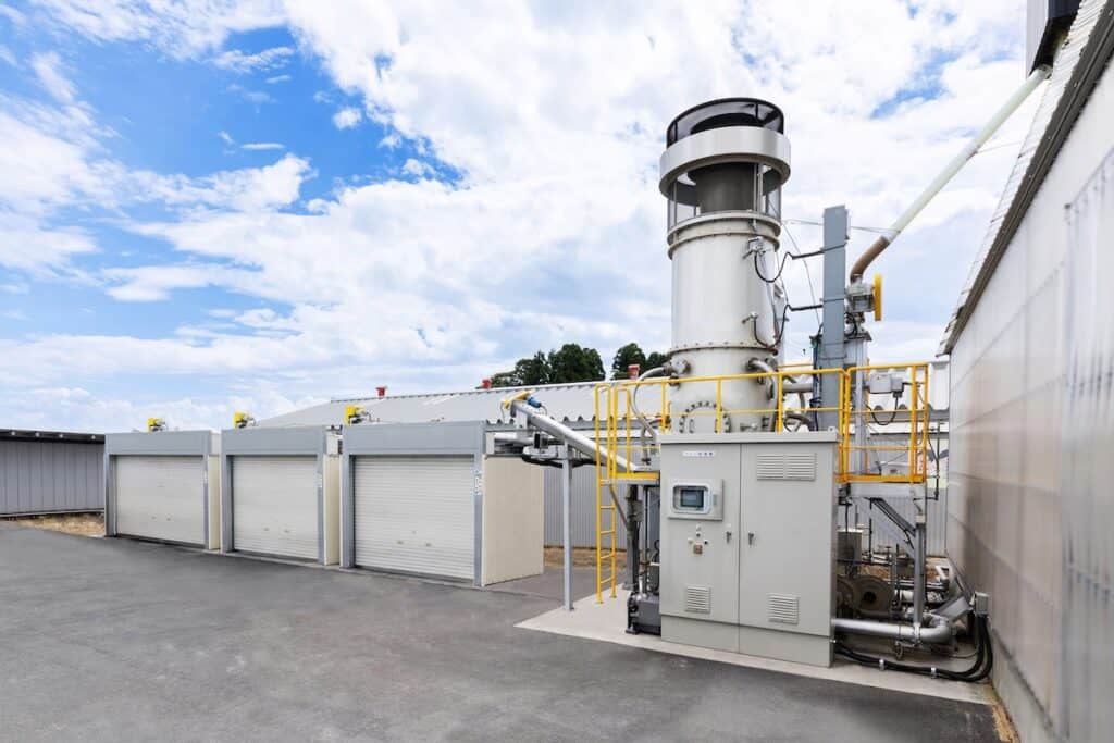 Rendering of Yanmar Rice Husk Biochar Production System with grey industrial plant alongside garage-like units in yard, blue skies behind.