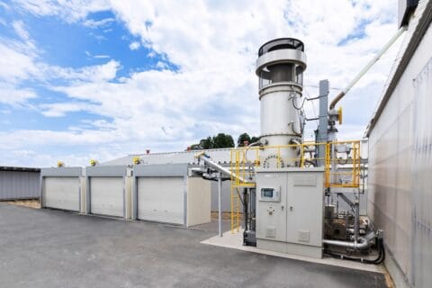 Rendering of Yanmar Rice Husk Biochar Production System with grey industrial plant alongside garage-like units in yard, blue skies behind.