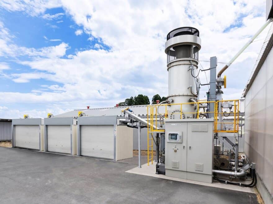 Rendering of Yanmar Rice Husk Biochar Production System with grey industrial plant alongside garage-like units in yard, blue skies behind.